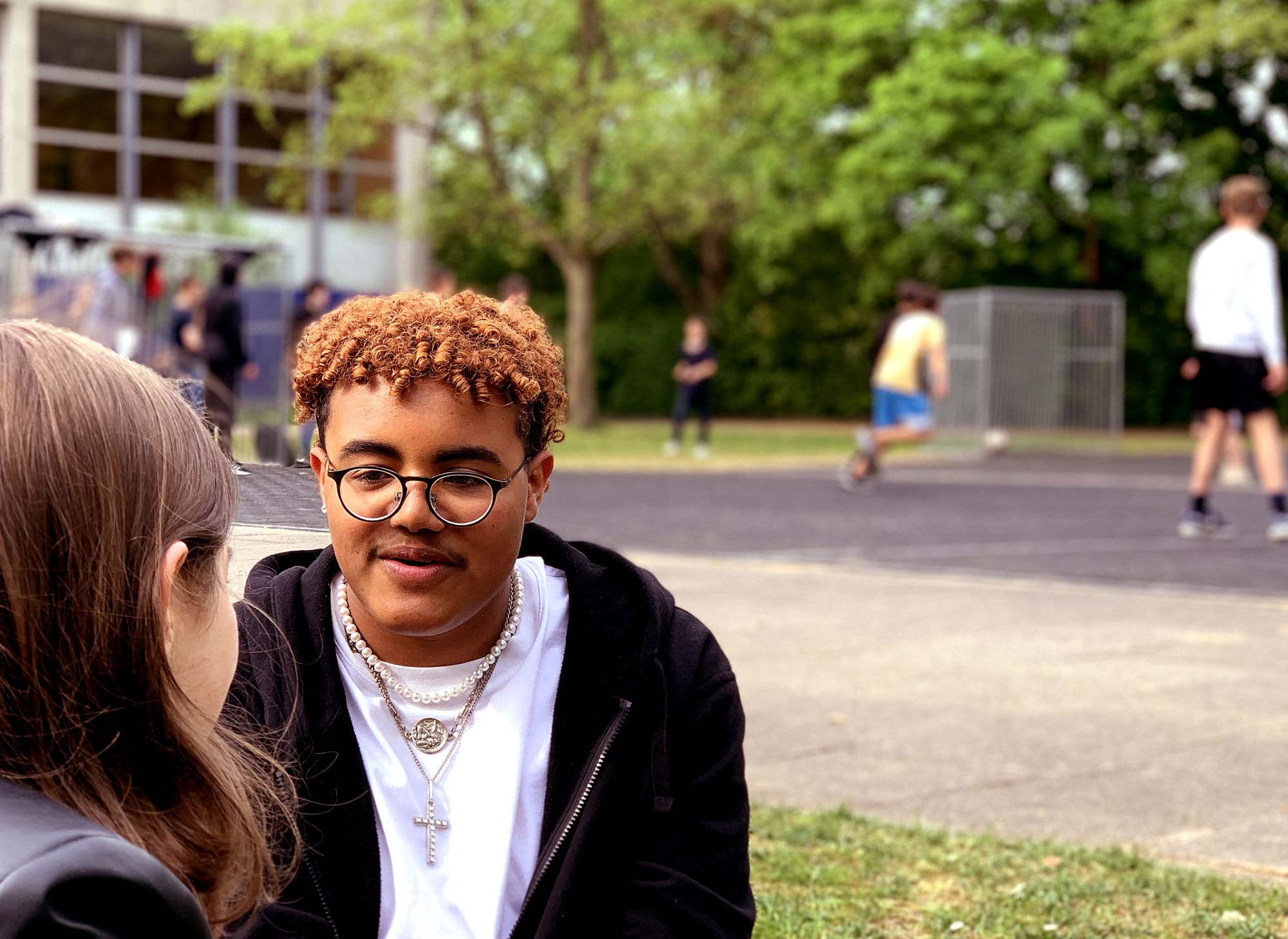Ein Junge unterhält sich mit einem Mädchen. Im Hintergrund ist unscharf ein Basketballplatz mit spielenden Jugendlichen zu sehen