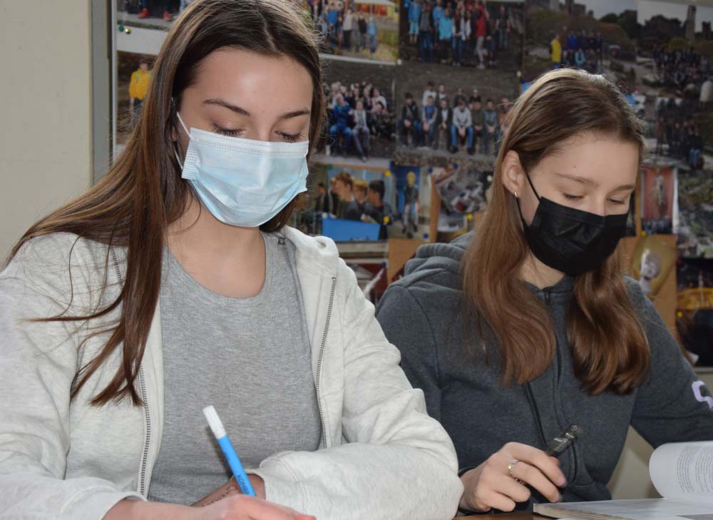 zwei Schülerinnen beim Arbeiten am Pult im Latein Raum. Im Hintergrund ist eine Fotowand aus dem Lateinkurs zu sehen