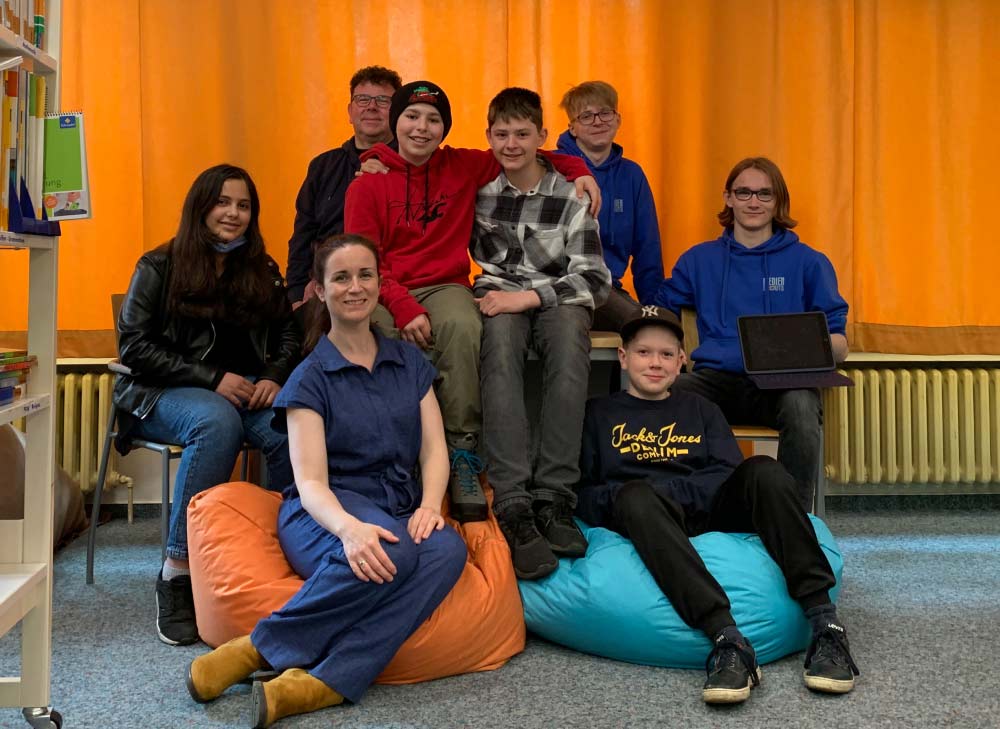 Gruppenbild der Medienscouts in der Schulbibliothek vor einem Fenster mit einem orangen Vorhang.