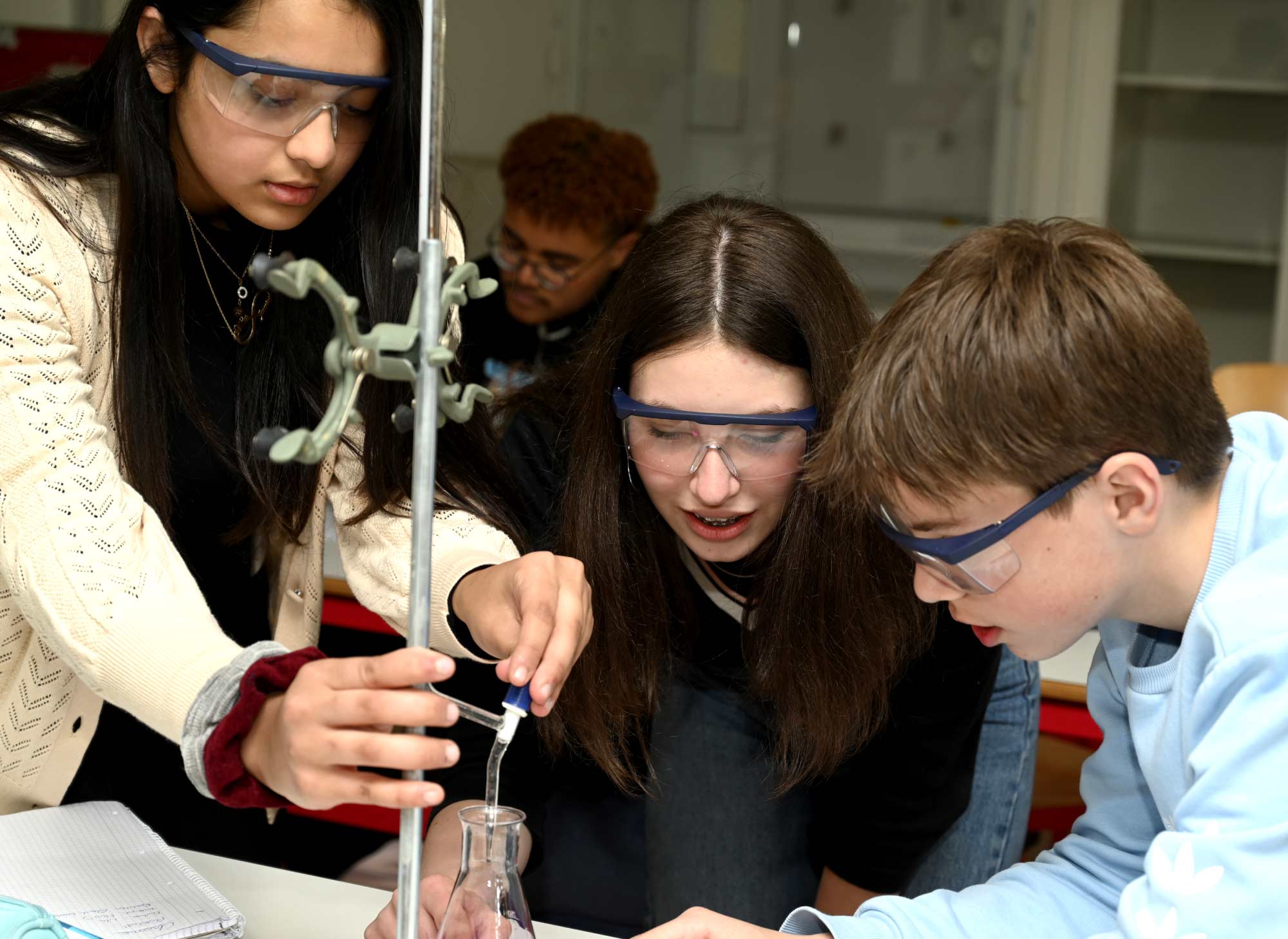 2 Mädchen ein Junge mit Labor-Schutzbrillen bei einem Chemischen Experiement. Ein Mädchen träufelt eine lila Flüssikeit in einem Kolben