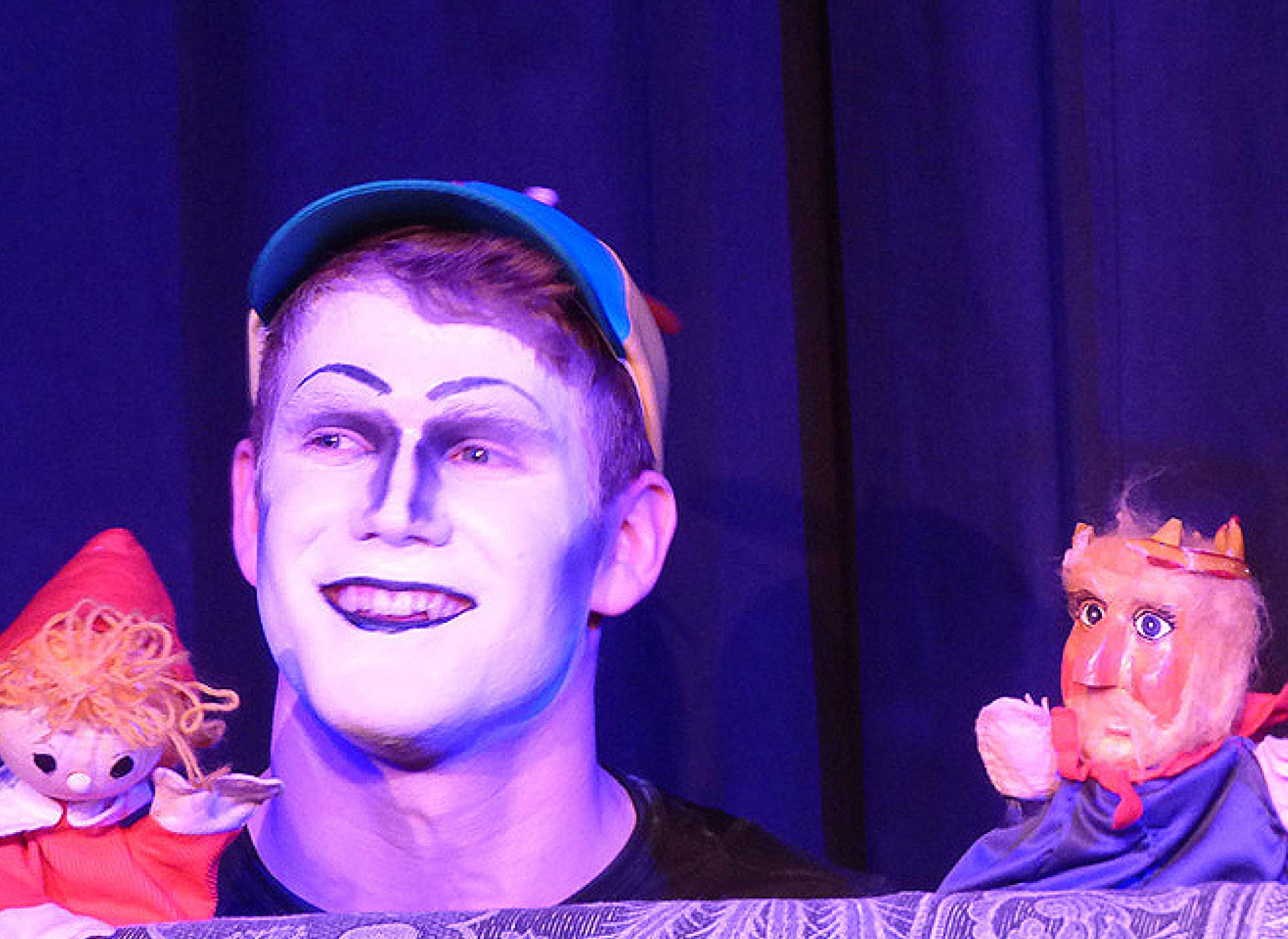 Aus einer Theatervorstellung: Ein Junge mit weiß geschminkten Gesicht und blau-gelber Kappe und zwei Kasperle-Figuren sind vor einem blauen Vorhang zu