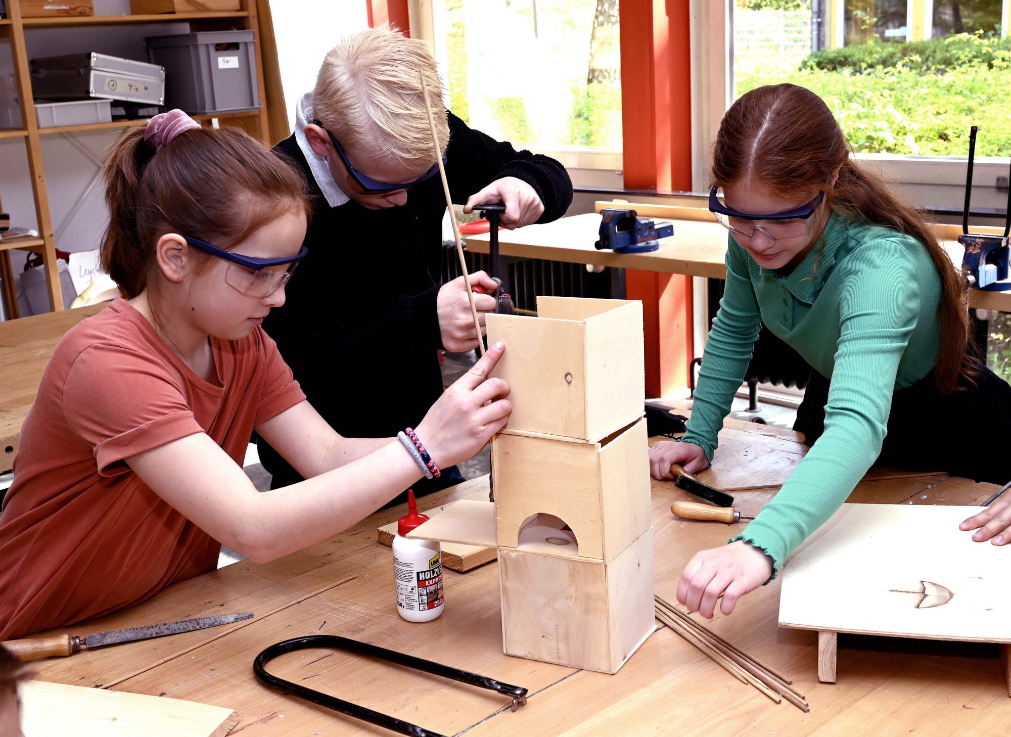 Zwei Schülerinnen und ein Schüler bauen an einem Holzkasten. Auf dem Holztisch sind ebenfalls Leim und weitere Wekzeuge und Materialien zu erkennen. I