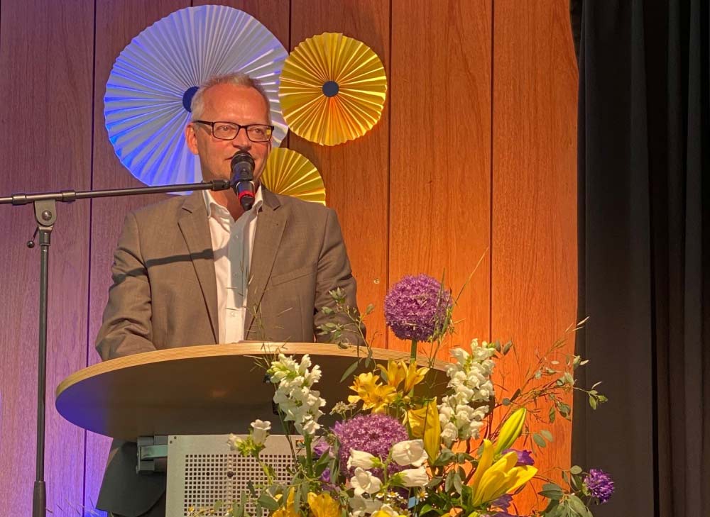 Schulleiter Sven Mérono bei seiner Rede am Rednerpult mit Lila, geln und weißen Blumen geschmückt auf einer orange beleuchteten Bühne 