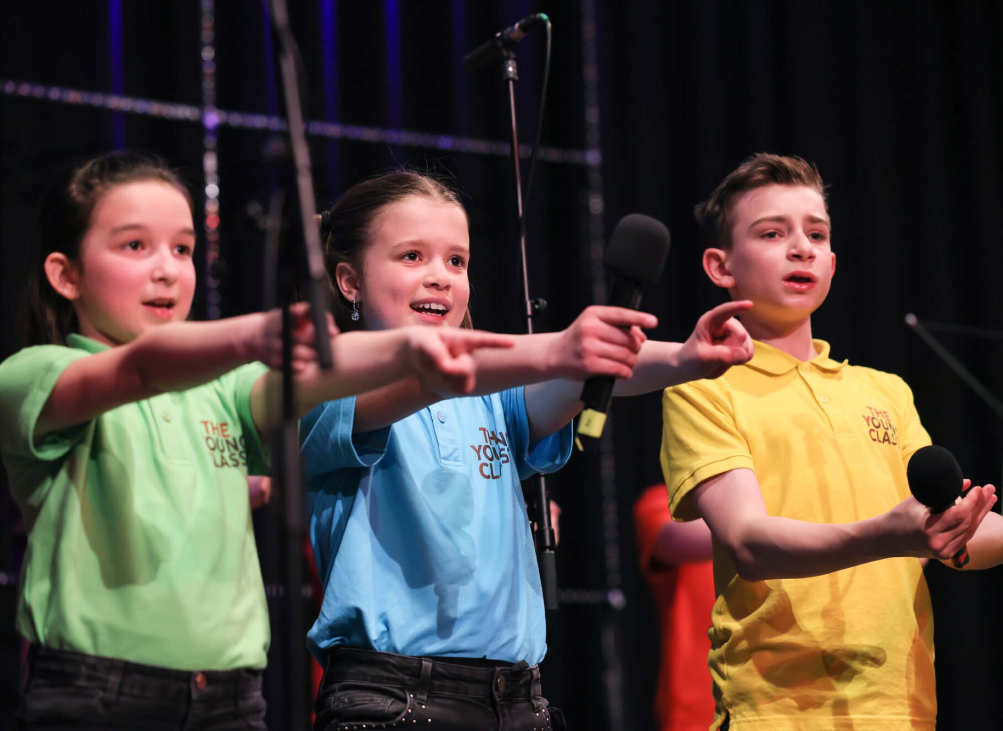 Schüler:innen in bunten T-Shirt in Nahaufnahme auf der Bühne zeigen mit beiden Armen ins Publikum und singen.