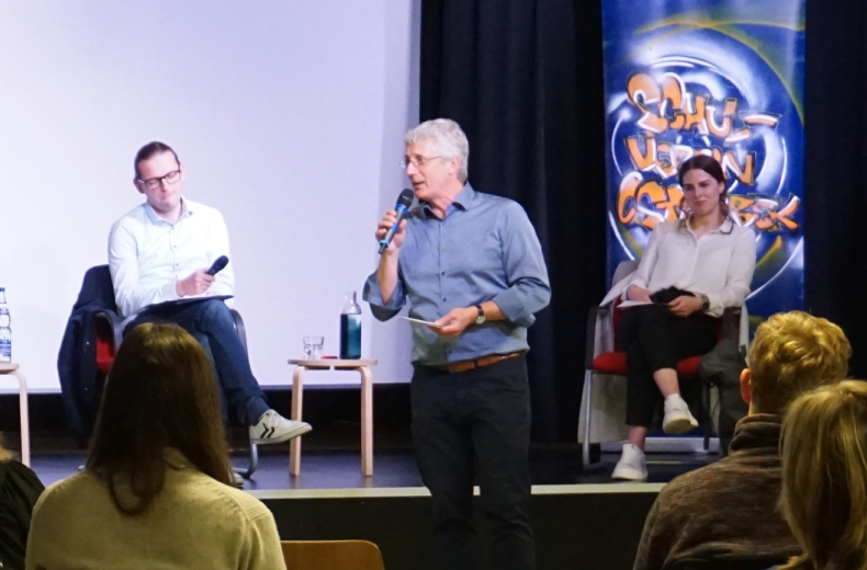 Drei Erwachsene auf einer Podeumsdiskussion auf einer Bühne mit Schülern als Publikum im Vordergrund